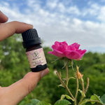 Flacone di olio essenziale di Rosa Damascena Aromabano tenuto in mano accanto a una rosa fresca.